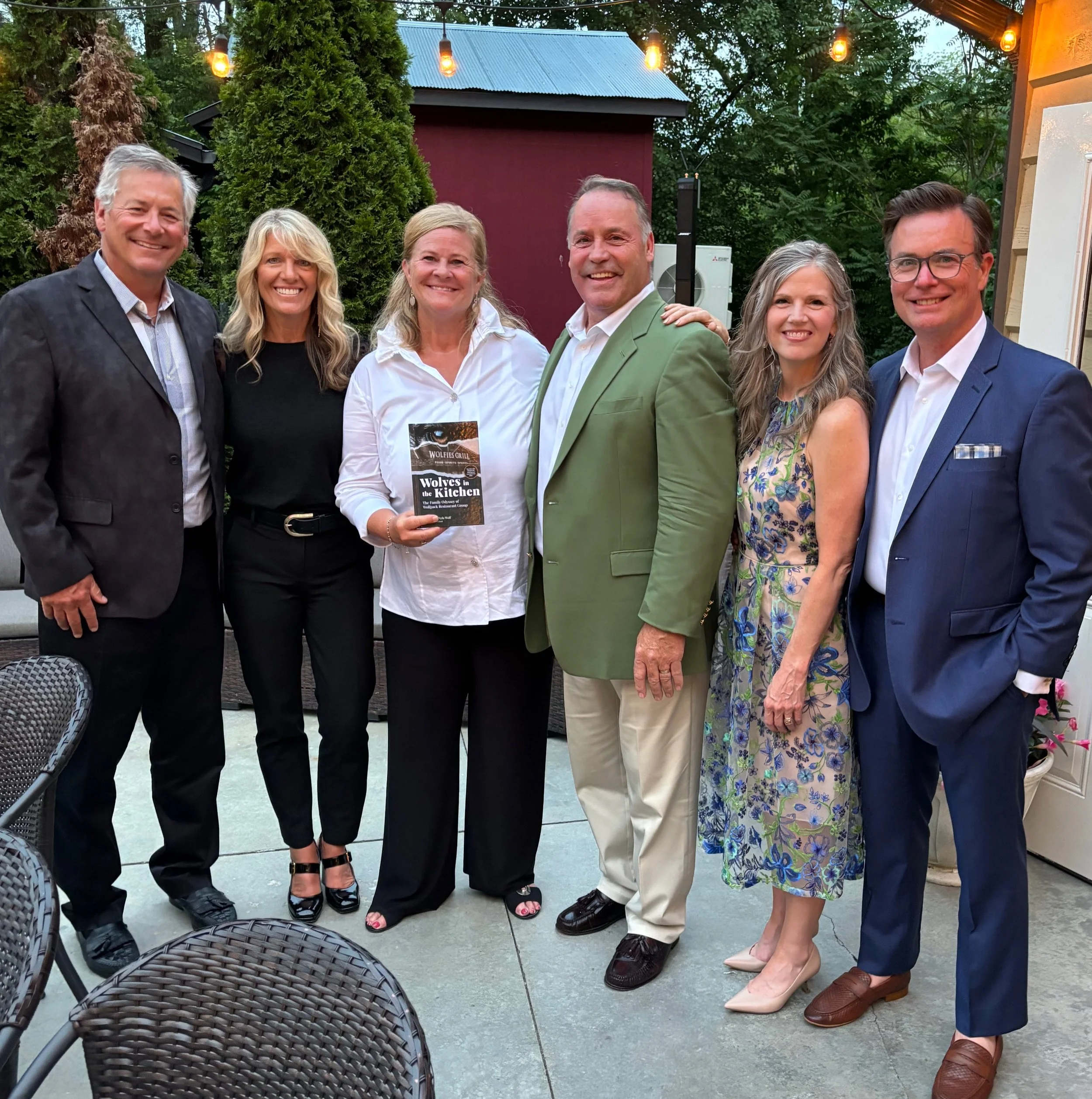 Emily & her husband Scott with clients at a book release party, smiling, with string lights overhead and a small red building in the background. Nyla Wolf holds a copy of the book  'Wolves in the Kitchen' that Emily helped Scott and Nyla Wolf write.