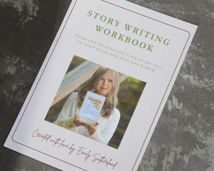 A booklet titled "Story Writing Workbook" with a subtitle about exercises to help get your story out of your head and into the world. The cover features a woman holding the workbook and standing outdoors, with a signature that reads "Created with love by Emily Sutheland." The booklet is placed on a textured gray surface.