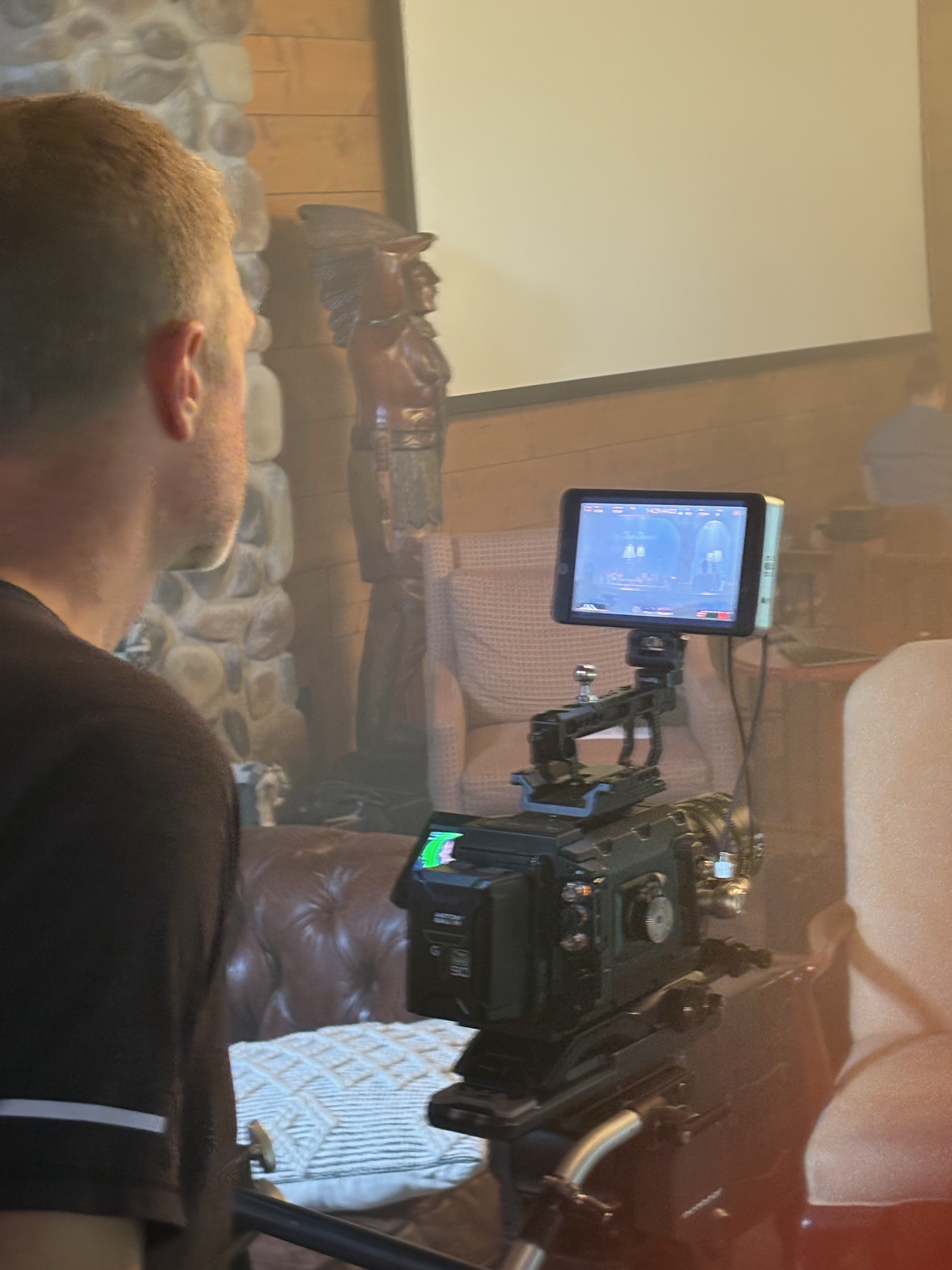 A man operating a camera on a tripod, with a film set or production area in the background, including a wooden wall and a carved wooden figure.