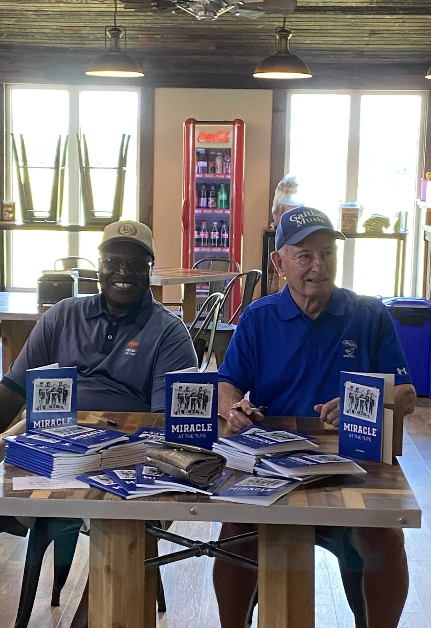 Two men sitting at a table with promotional materials for a book titled 'Miracle at the Tute'. The man on the left is smiling and wearing glasses, a tan cap, and a dark polo shirt. The man on the right is holding a pen and wearing a blue shirt and a blue cap. In the background, there are windows, a Coca-Cola refrigerator, and some shelves.