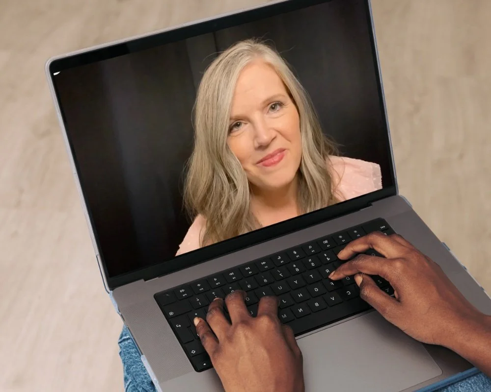 A person with dark skin typing on a laptop keyboard, which displays a woman with blonde hair and a pink top on the screen.