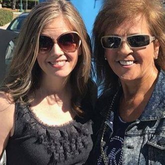 Two women smiling for a photo outdoors, one younger with long blonde hair wearing sunglasses and a black top, and one older with short light brown hair wearing sunglasses and a denim jacket.