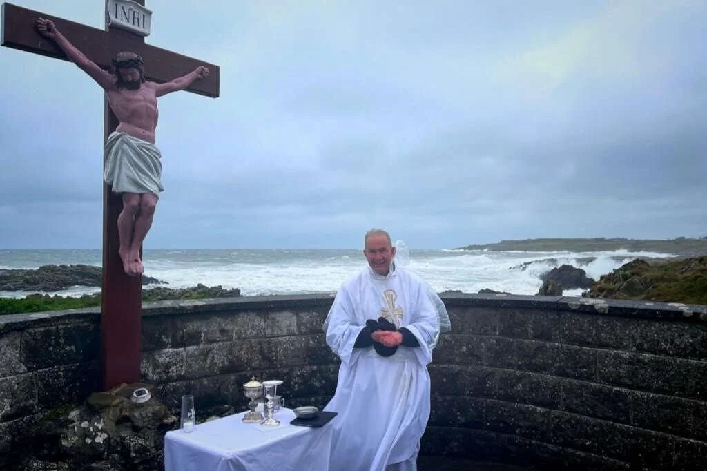 Fr Gerry McCloskey beside an outdoor altar and coastal crucifix
