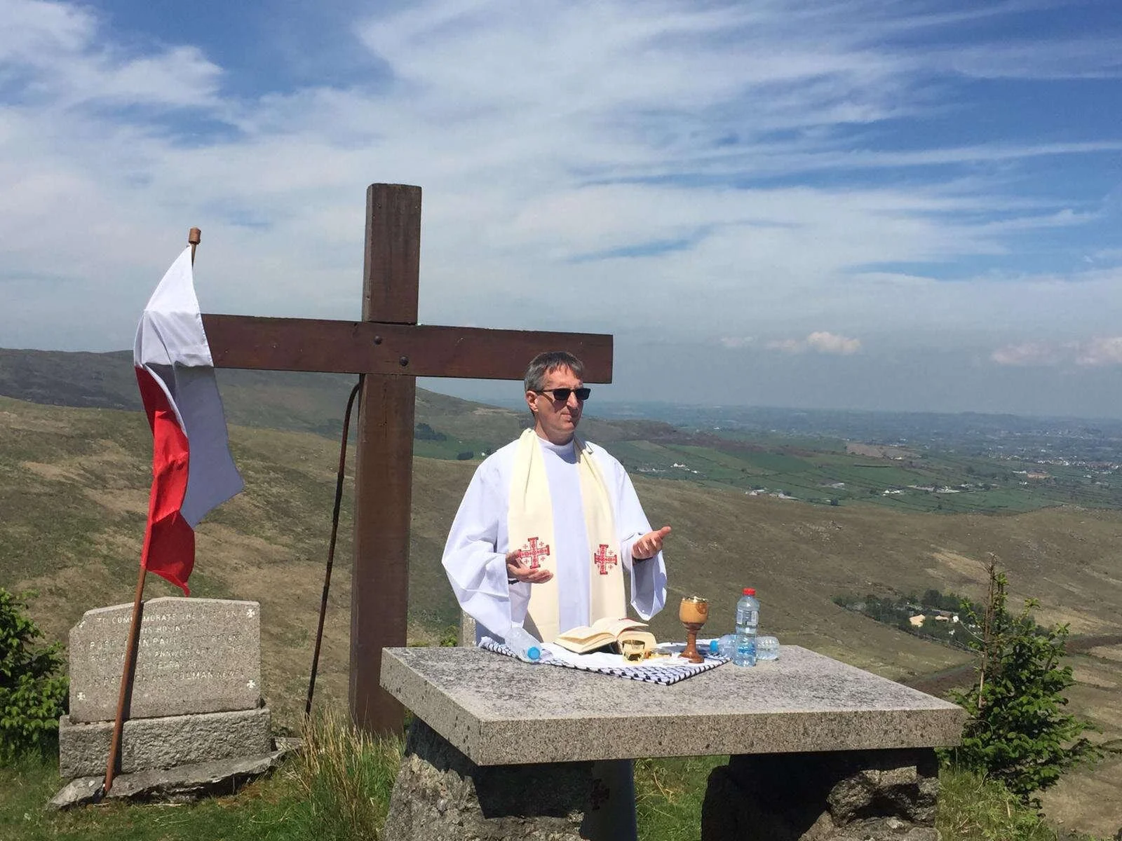 A priest celebrates Mass beside a cross and tricolour on a hillside