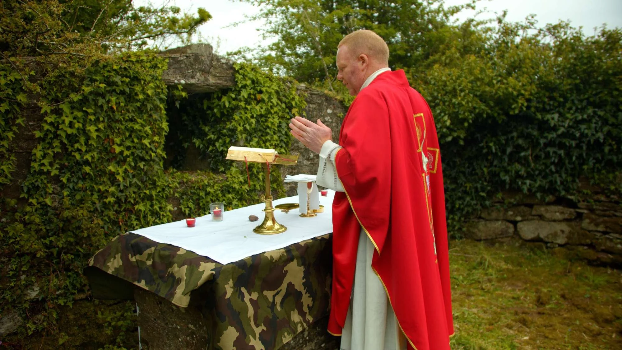 A priest prays at an outdoor altar wearing red vestments
