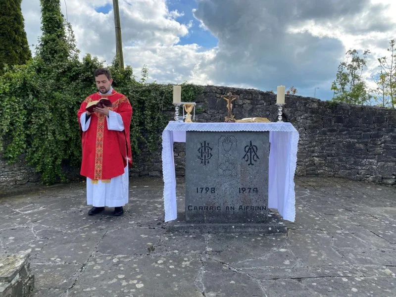 A priest stands beside an inscribed Mass Rock altar