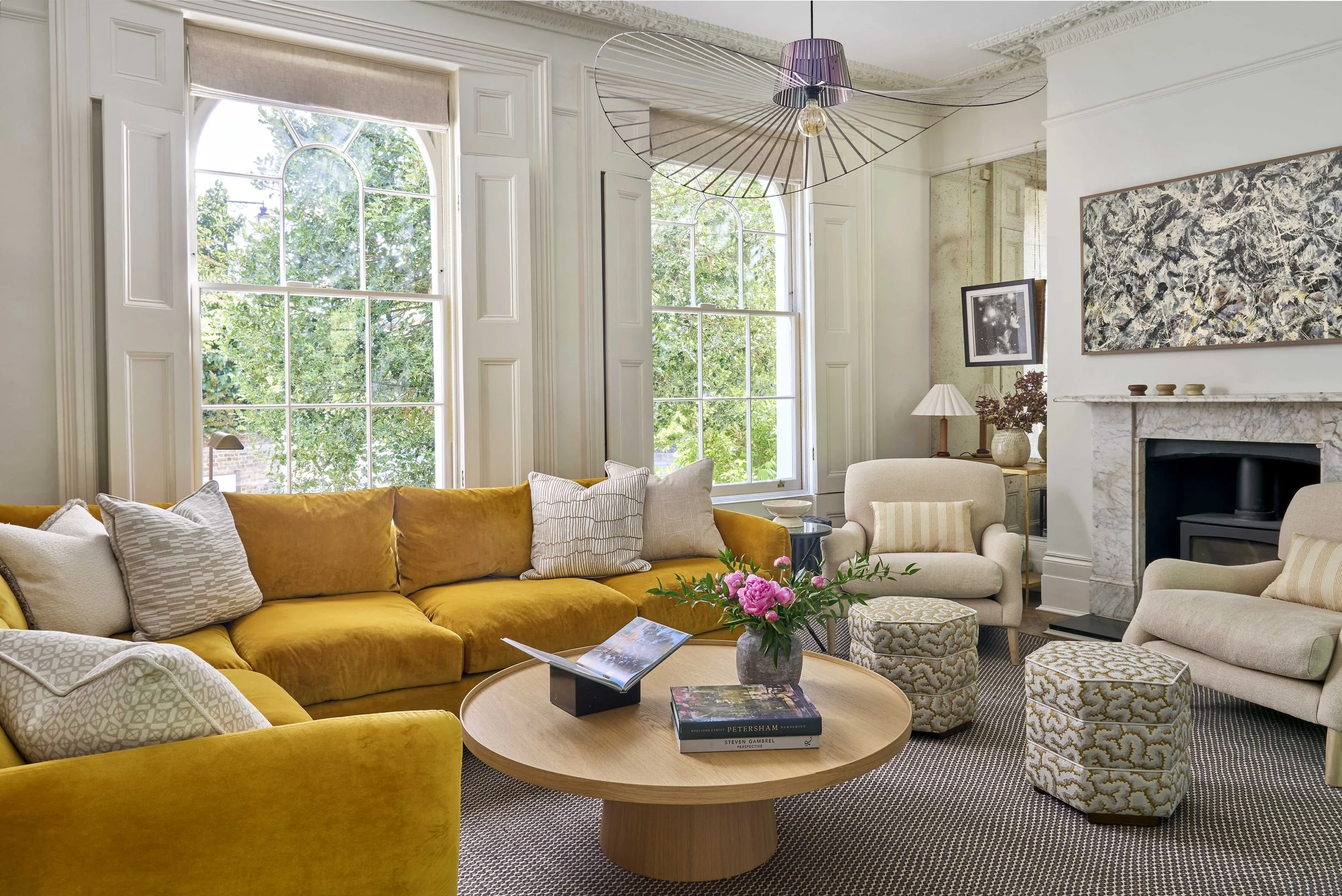 A living room with large windows, a yellow velvet sofa, a round wooden coffee table with books and a vase, beige armchairs, a marble fireplace, and wall art, with natural light coming through the windows. Collected and curated interior in London.