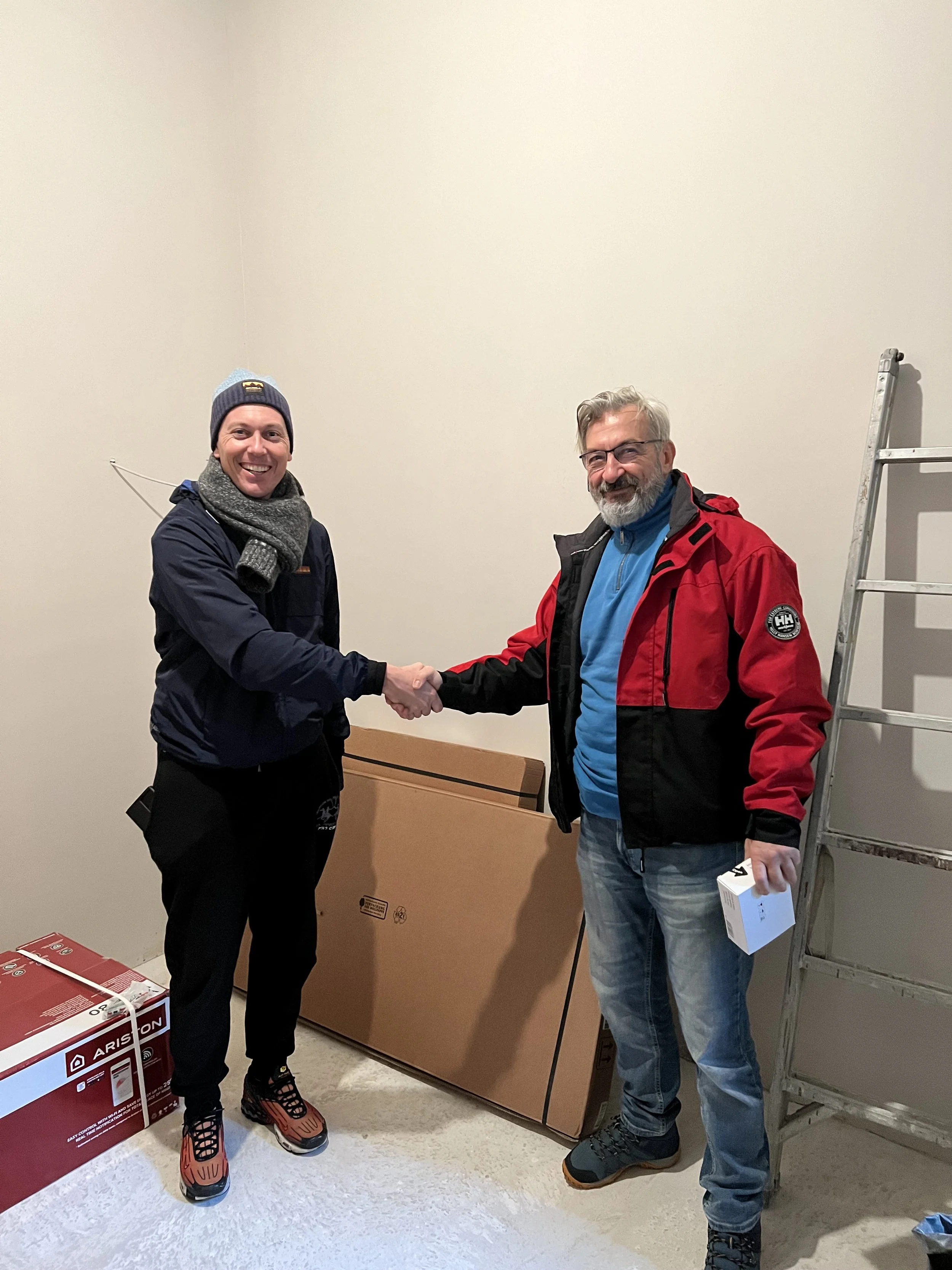 Two men shake hands in a room with boxes and a ladder, one is wearing a beanie, scarf, and winter jacket, the other has a beard, glasses, and a red and black jacket.
