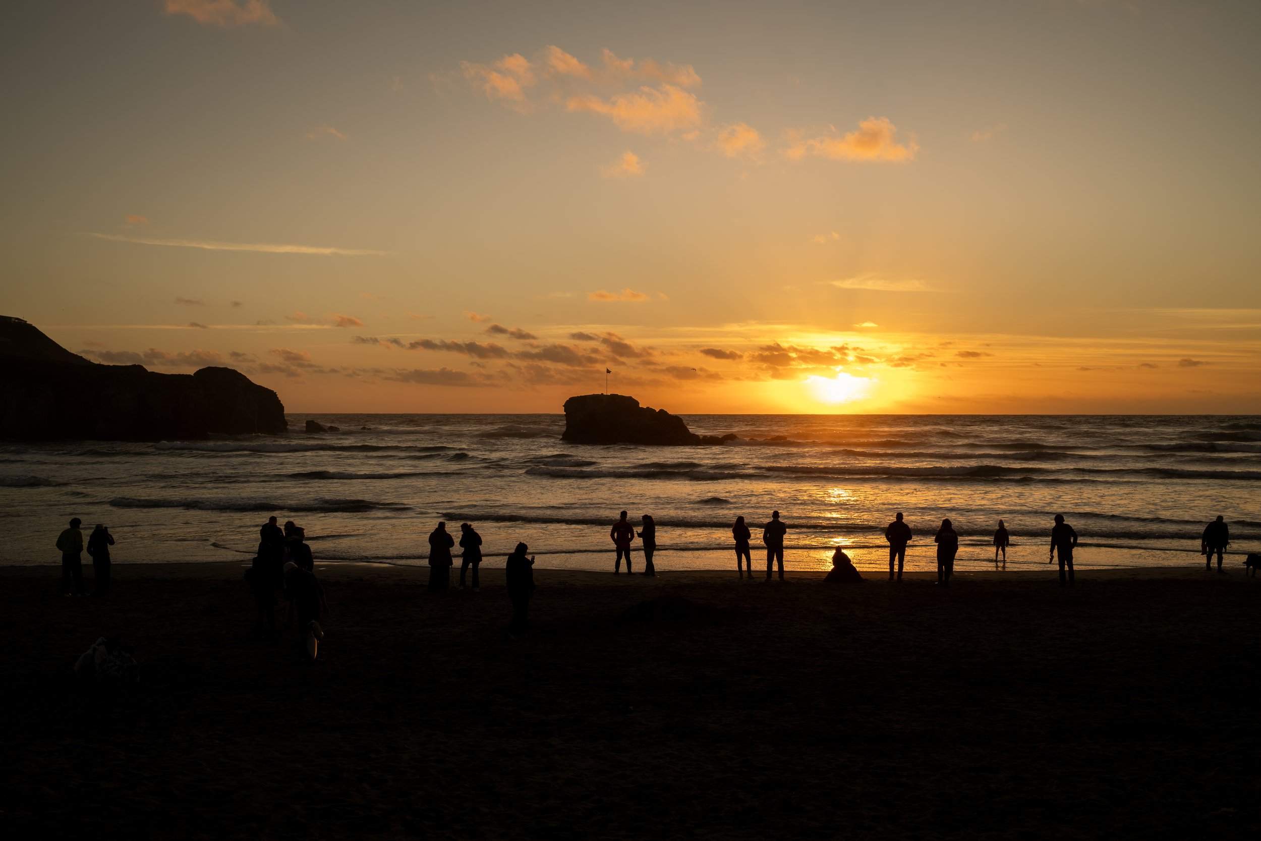 People silhouetted on a beach during sunset, with the sun near the horizon, a rock formation in the ocean with a flag on top, and a sky with scattered clouds.