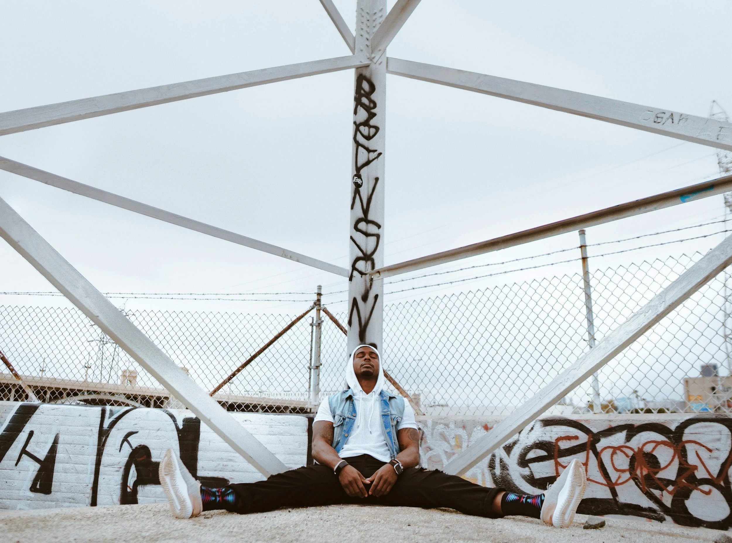 A person of colour sitting against a frame with graffiti behind them. Wearing a white short sleeve hoodie, denim vest and black pants, colourful socks and sneakers. Eyes closed, looking relaxed.