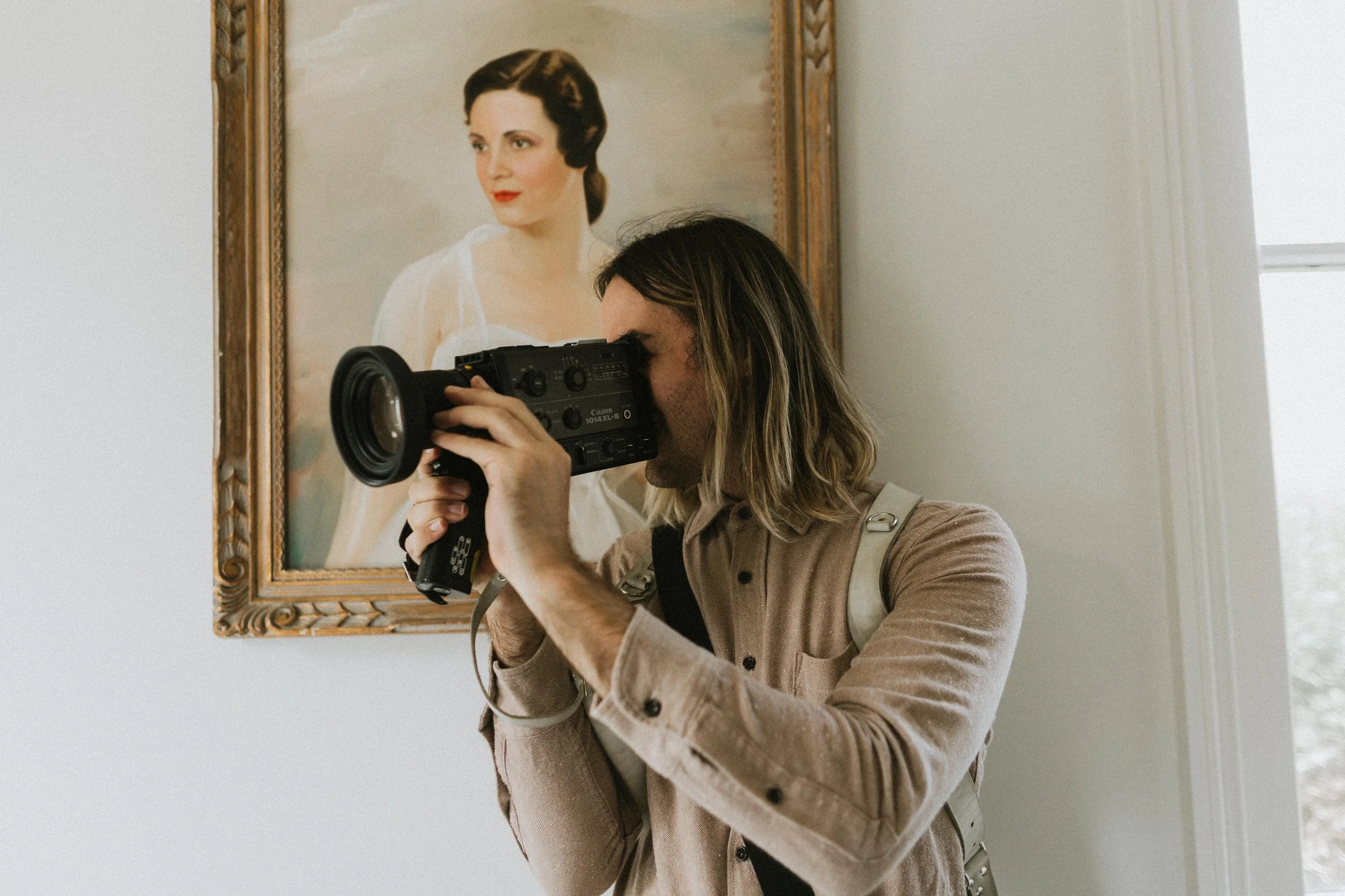 Photographer with long hair and a backpack takes a picture of a portrait of a woman with dark hair styled in vintage waves, red lipstick, and a white dress, hanging on a wall near a window.