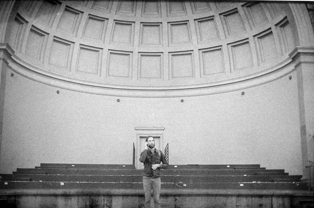 A man standing on a stage with a domed wall behind him, inside a large auditorium or theater.