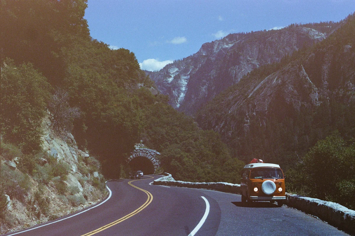 A vintage orange Volkswagen van driving along a winding mountain road surrounded by tall rocky mountains and greenery, with a distant tunnel entrance in the background under a partly cloudy sky.