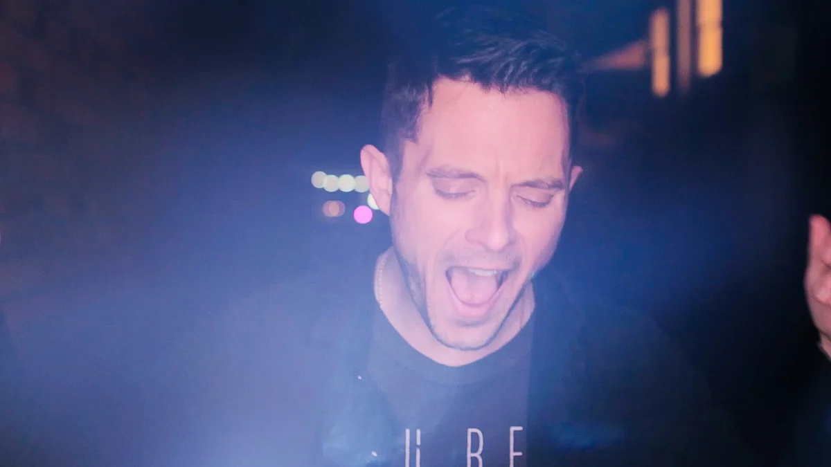 A man with dark hair and light skin, wearing a black shirt, appears to be singing or shouting with his eyes closed in a dark setting with some blurred lights in the background.