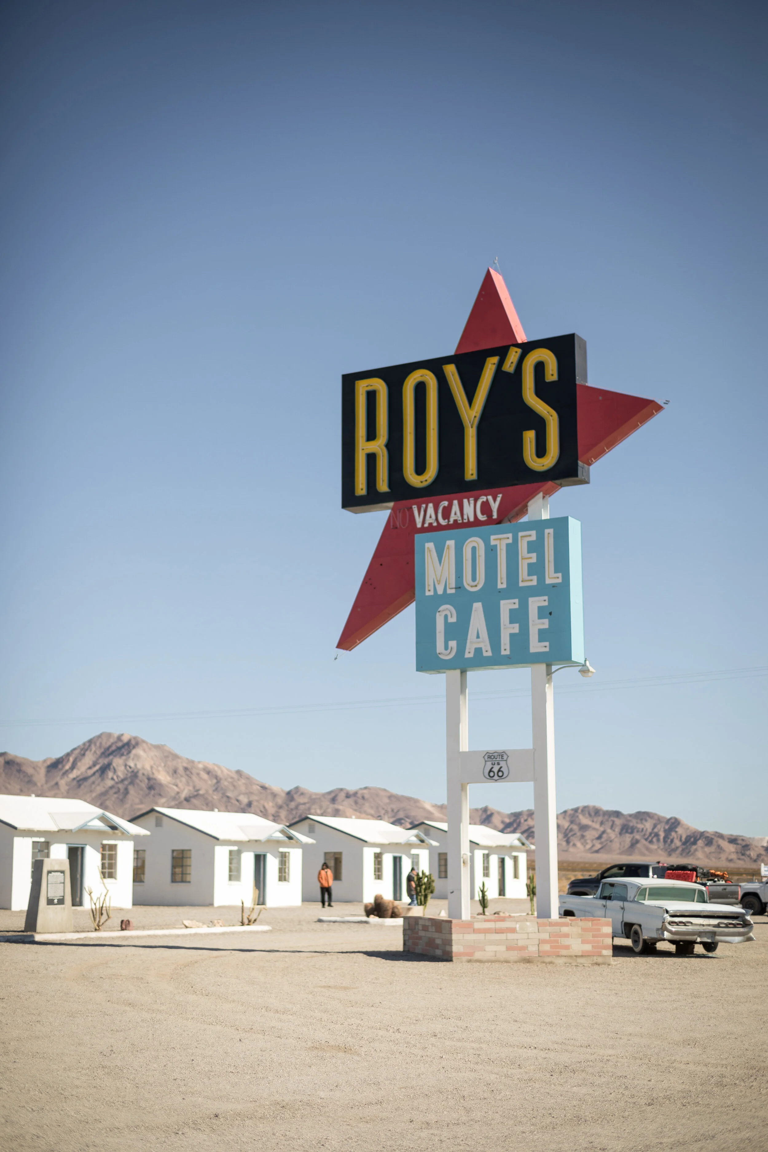 A vintage roadside sign advertising Roy's Motel and Café with a star design, set against a desert landscape with mountains and a row of white cottages.
