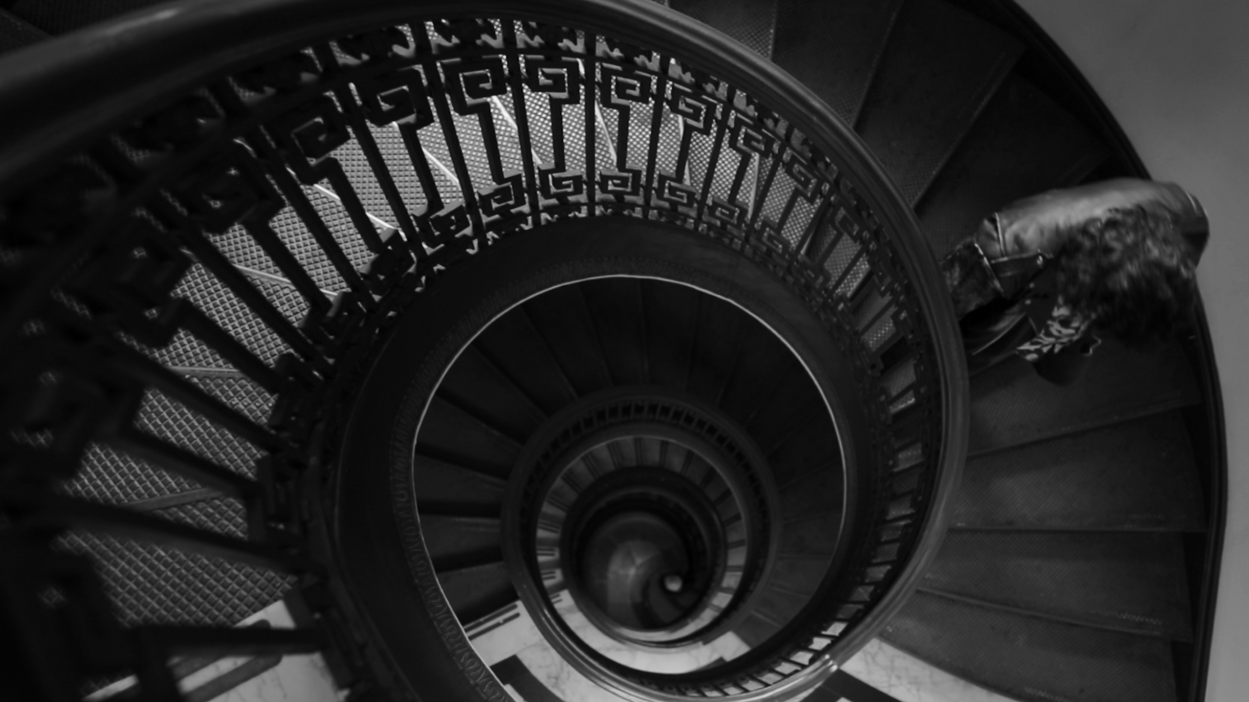 Black and white photo of a spiral staircase with a decorative railing, viewed from above, with a woman walking down the stairs.