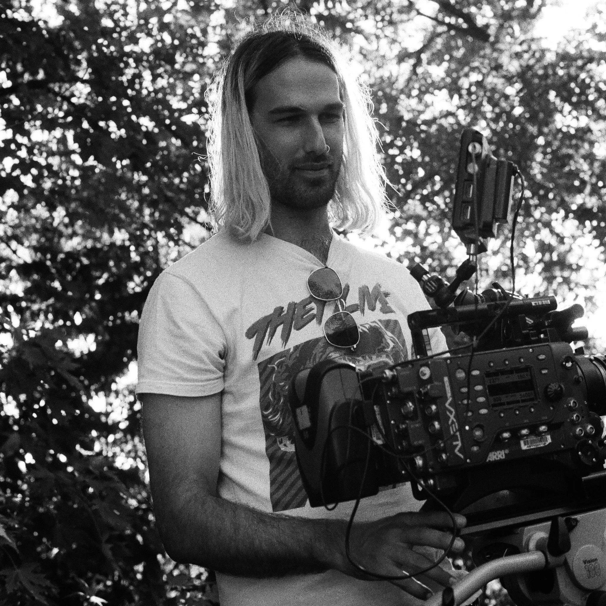 A man with long hair and a beard holding a camera rig outdoors, with trees and sunlight in the background.