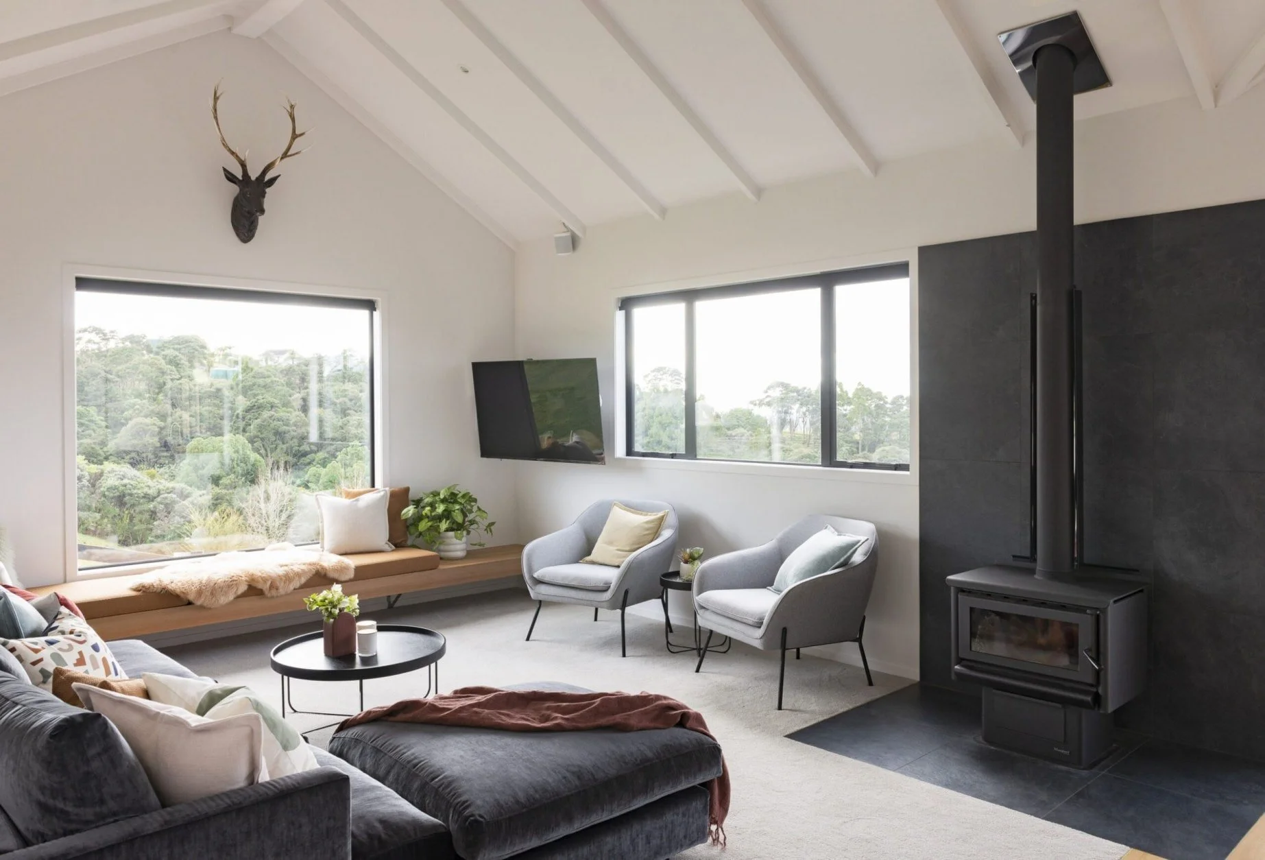 Modern country living room with vaulted ceilings, wood-burning fireplace, built-in window seat, and soft grey furnishings overlooking lush Clevedon bush views.