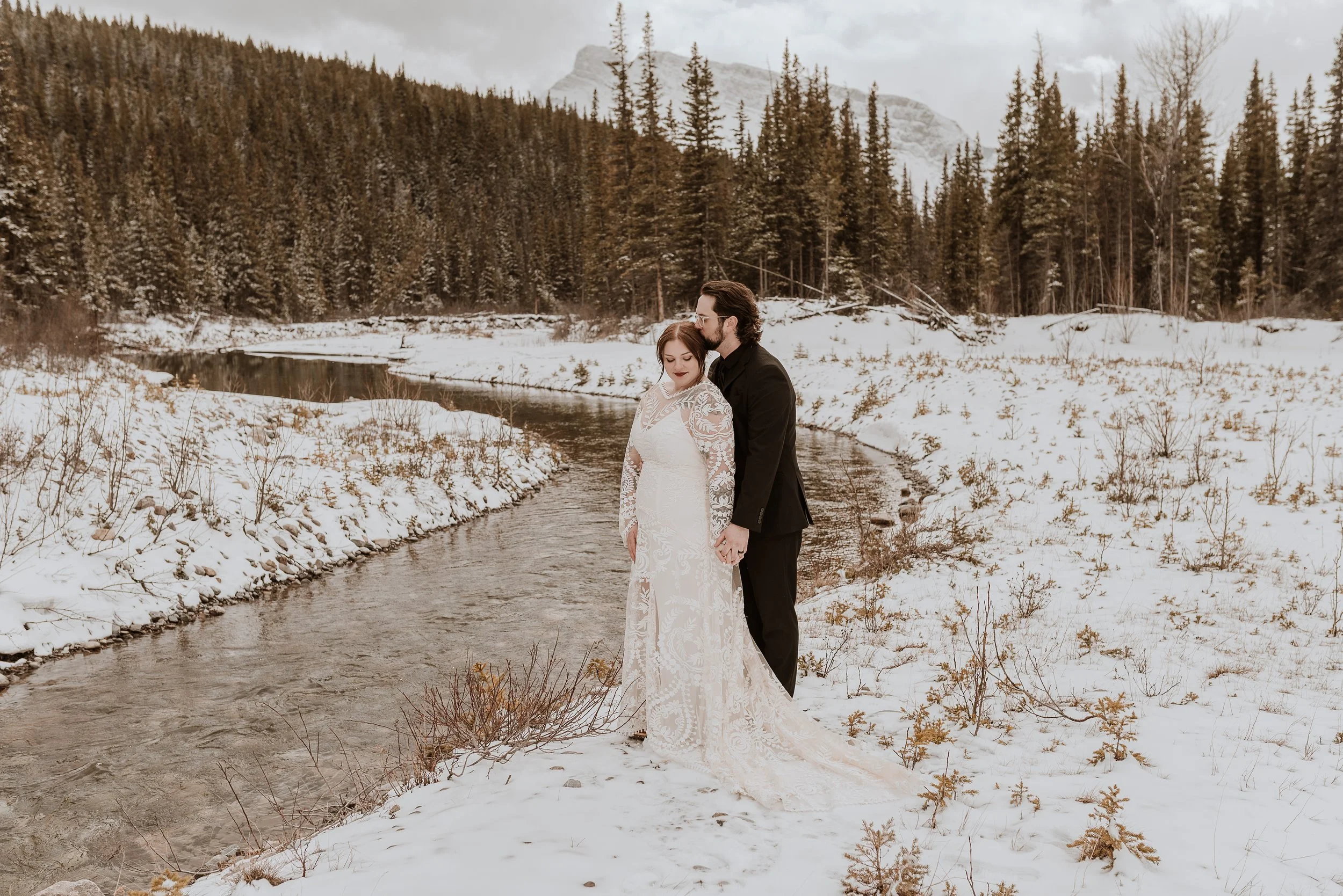 Winter wedding Alberta Canmore | Couple standing by stream snow on the ground