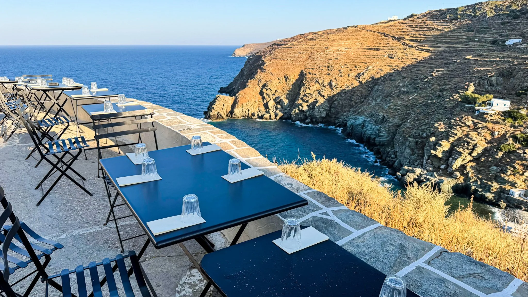 Loggia bar in Sifnos, Greece, with outdoor tables overlooking the Aegean Sea and rocky coastline in the golden evening light.