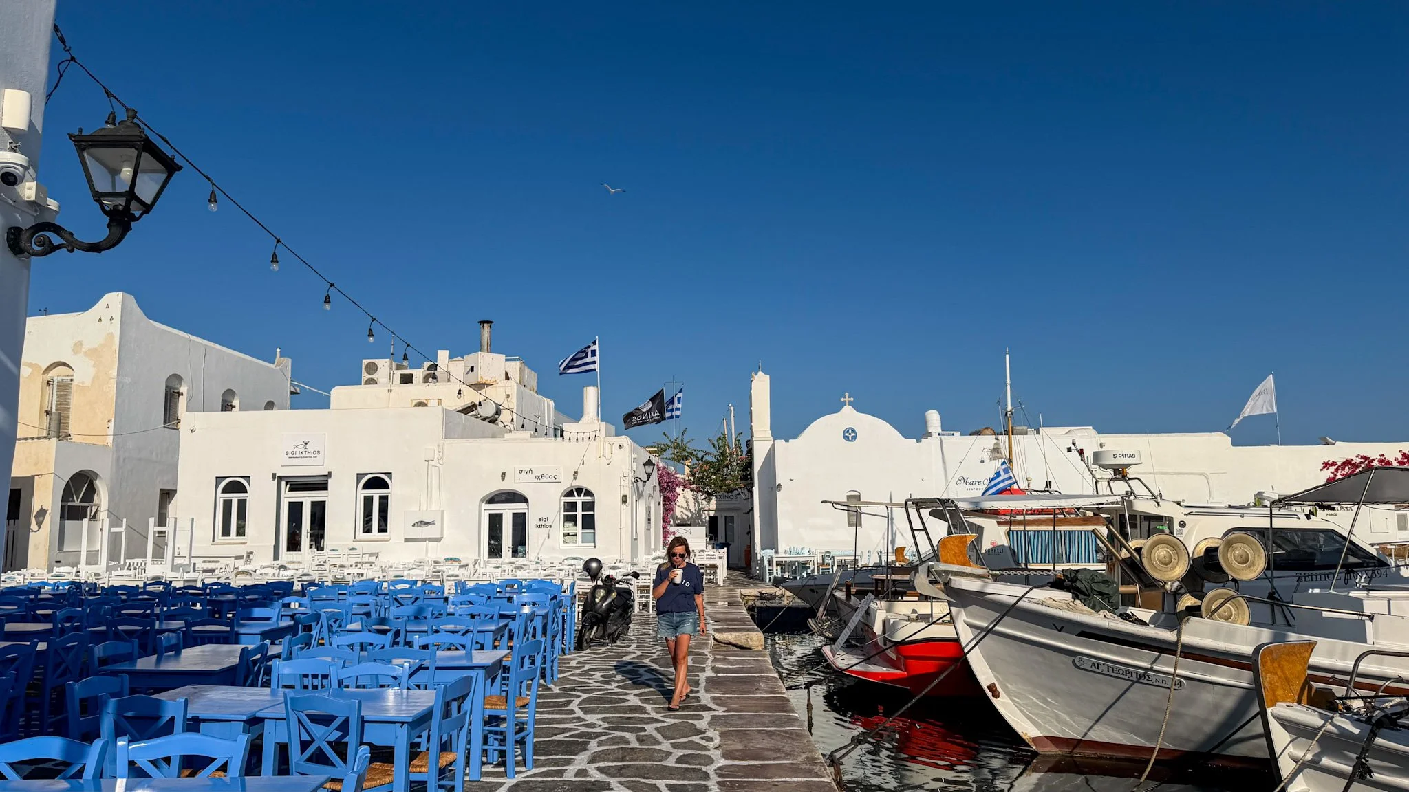 Naoussa harbour in Paros, Greece, with blue chairs at Barbarossa, whitewashed buildings and fishing boats along the waterfront.