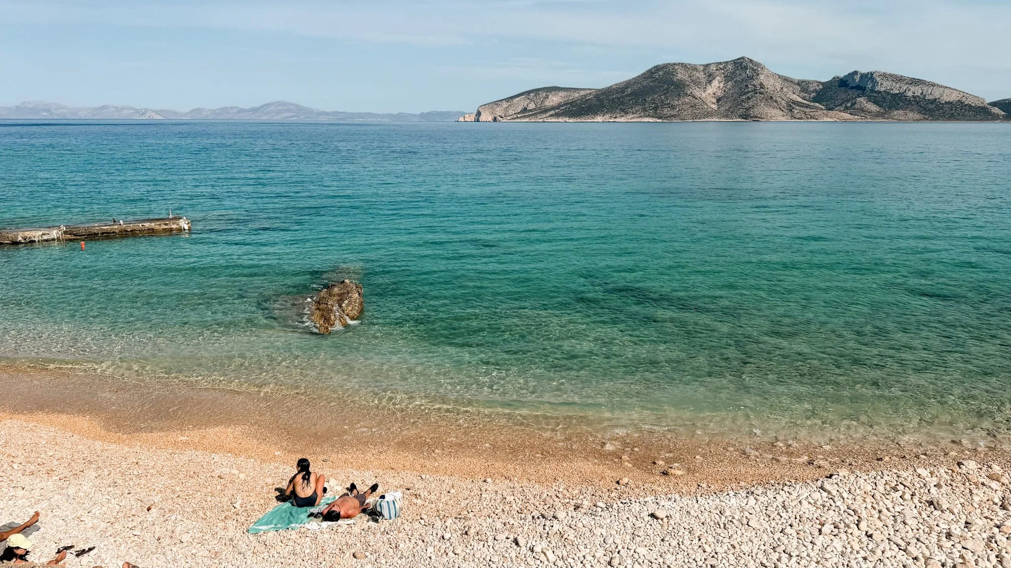 Crystal-clear turquoise water and a pebble beach in Koufonissia, Greece, with people sunbathing and views across the Aegean Sea.