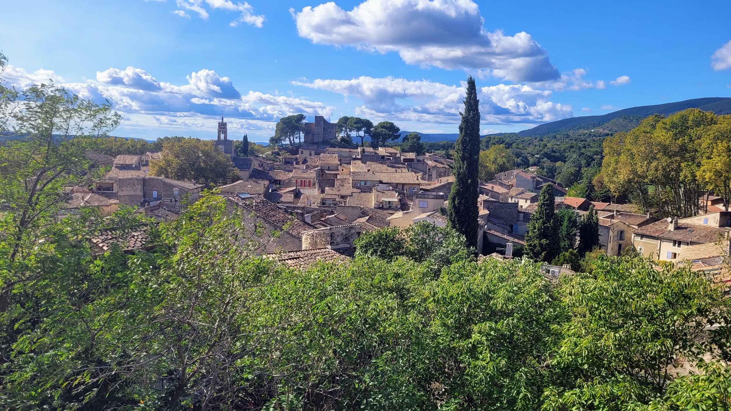View of a provençal town with tiled roofs surrounded by green trees and hills under a partly cloudy blue sky.