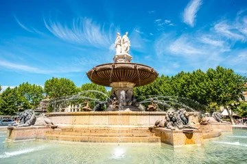 Central fountain with sculptures, water flowing, surrounded by greenery, under a blue sky with clouds.