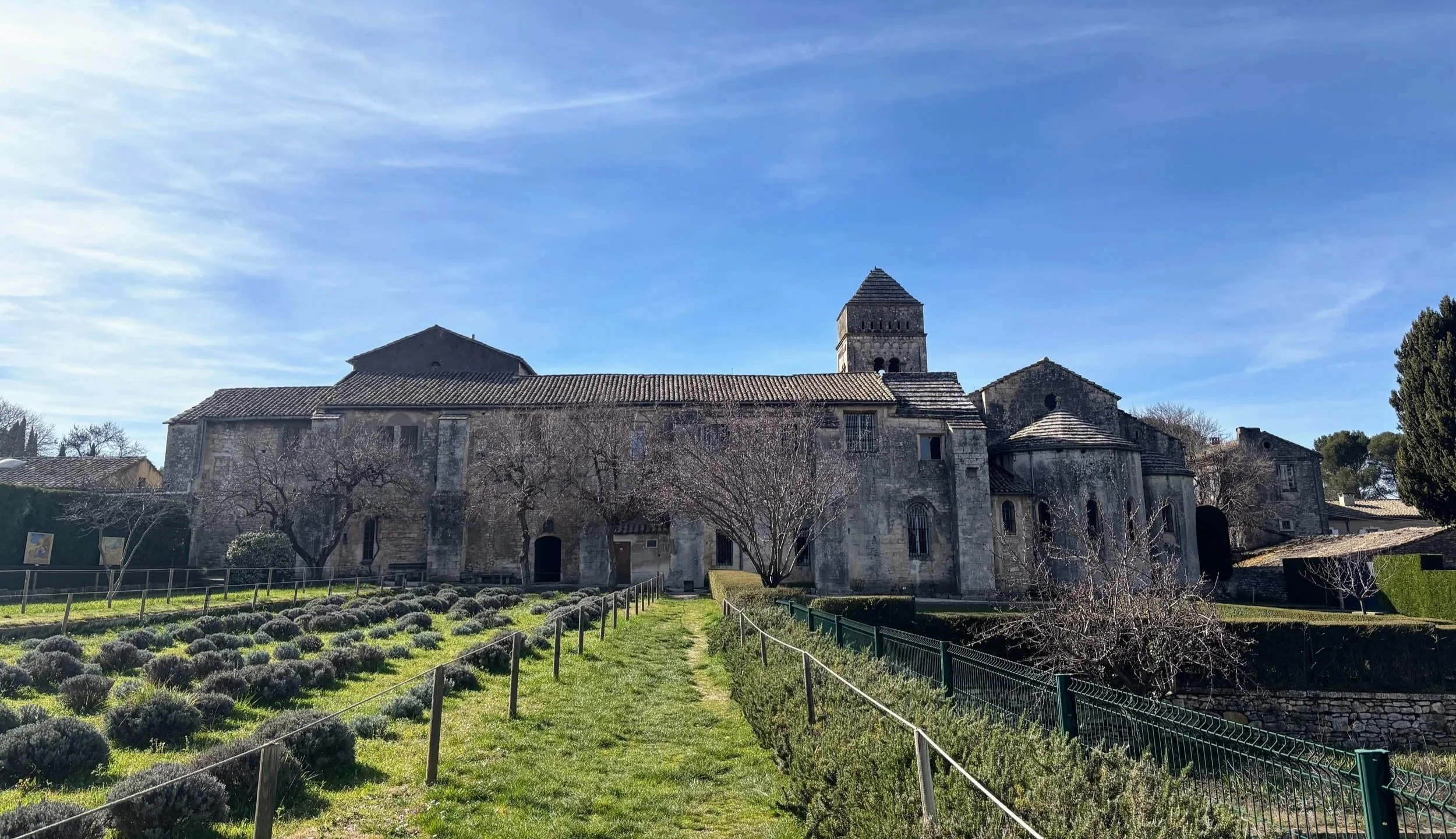 A historic stone church with a bell tower, surrounded by a well-maintained garden with lavender bushes and leafless trees, under a clear blue sky.