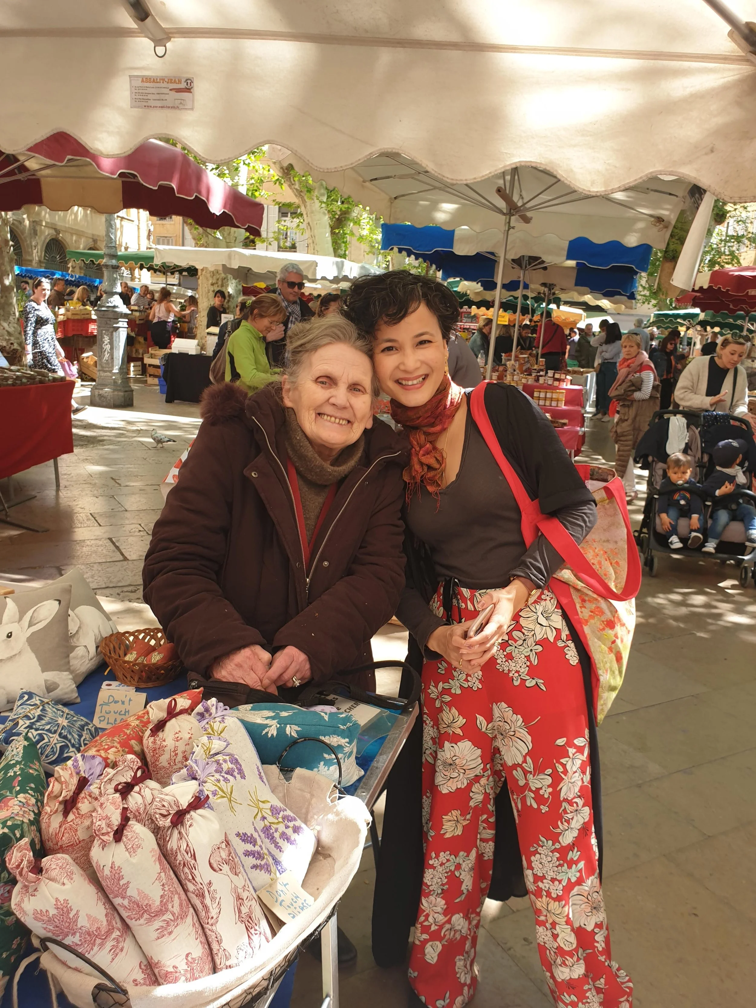 Two women smiling and standing close together at an outdoor marketplace with market stalls and other shoppers in the background.