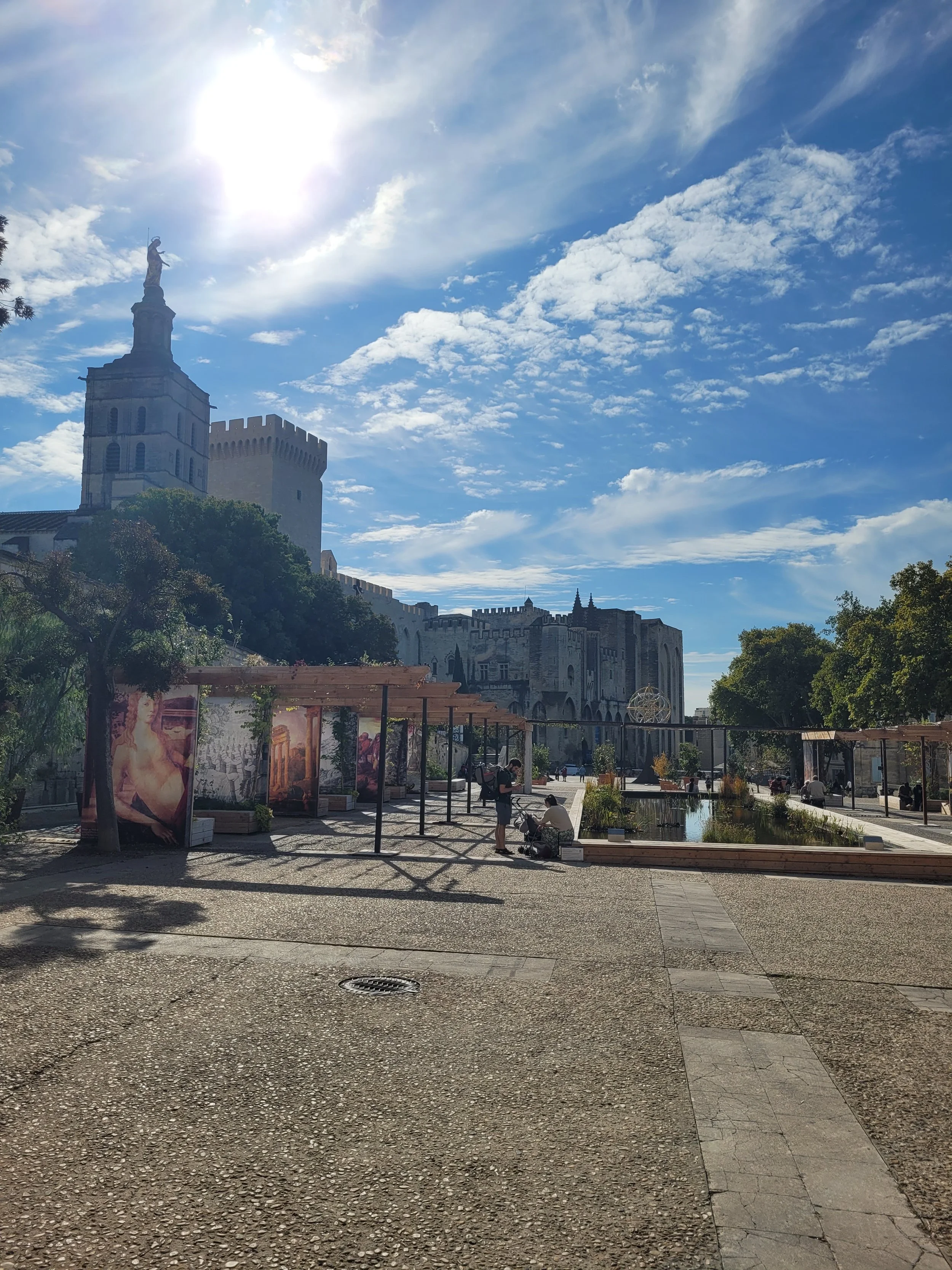 A historic castle with a tower and sculpture on top sits on a hill. The castle has crenellated walls and windows, set against a bright, partly cloudy sky with the sun shining.