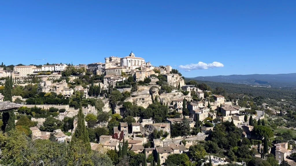 Gordes, A hillside town with densely packed stone buildings and lush greenery under a clear blue sky.