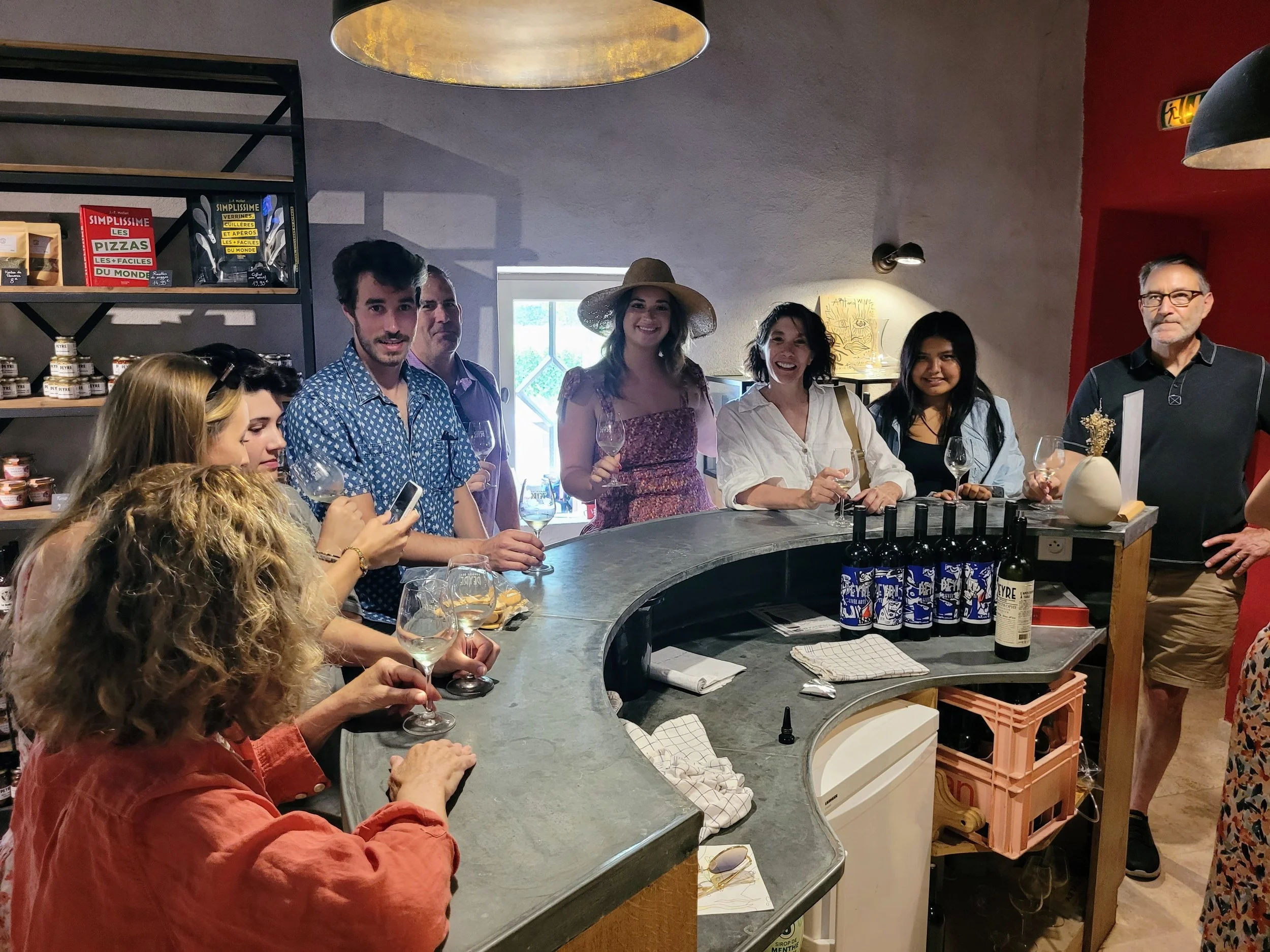 Group of people at a wine tasting event, some holding wine glasses, with various bottles and food items on the counter, in a cozy winery.
