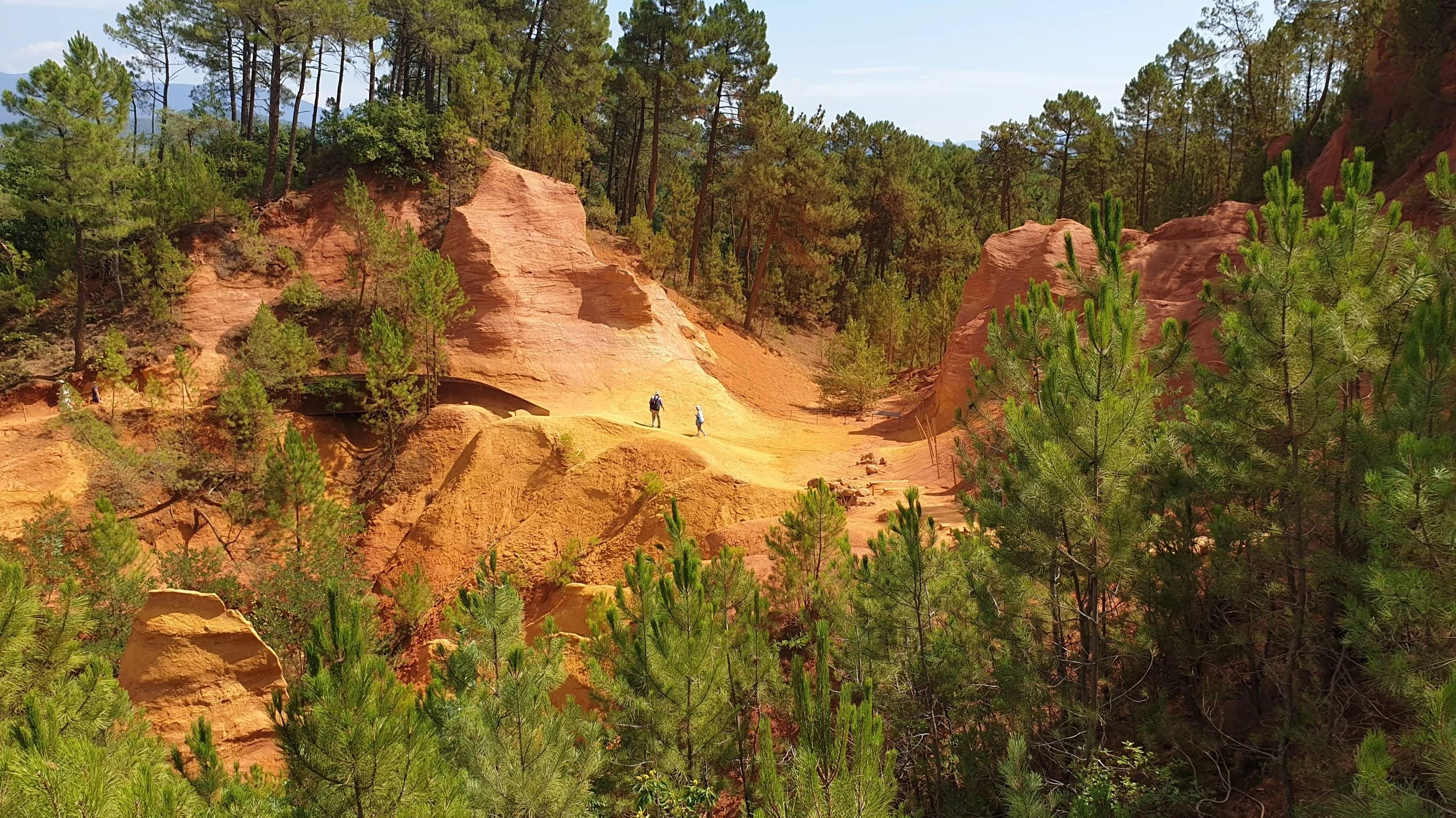 Red ochre formations and sandy trail in a desert landscape with green pine trees and three hikers walking among the rocks.
