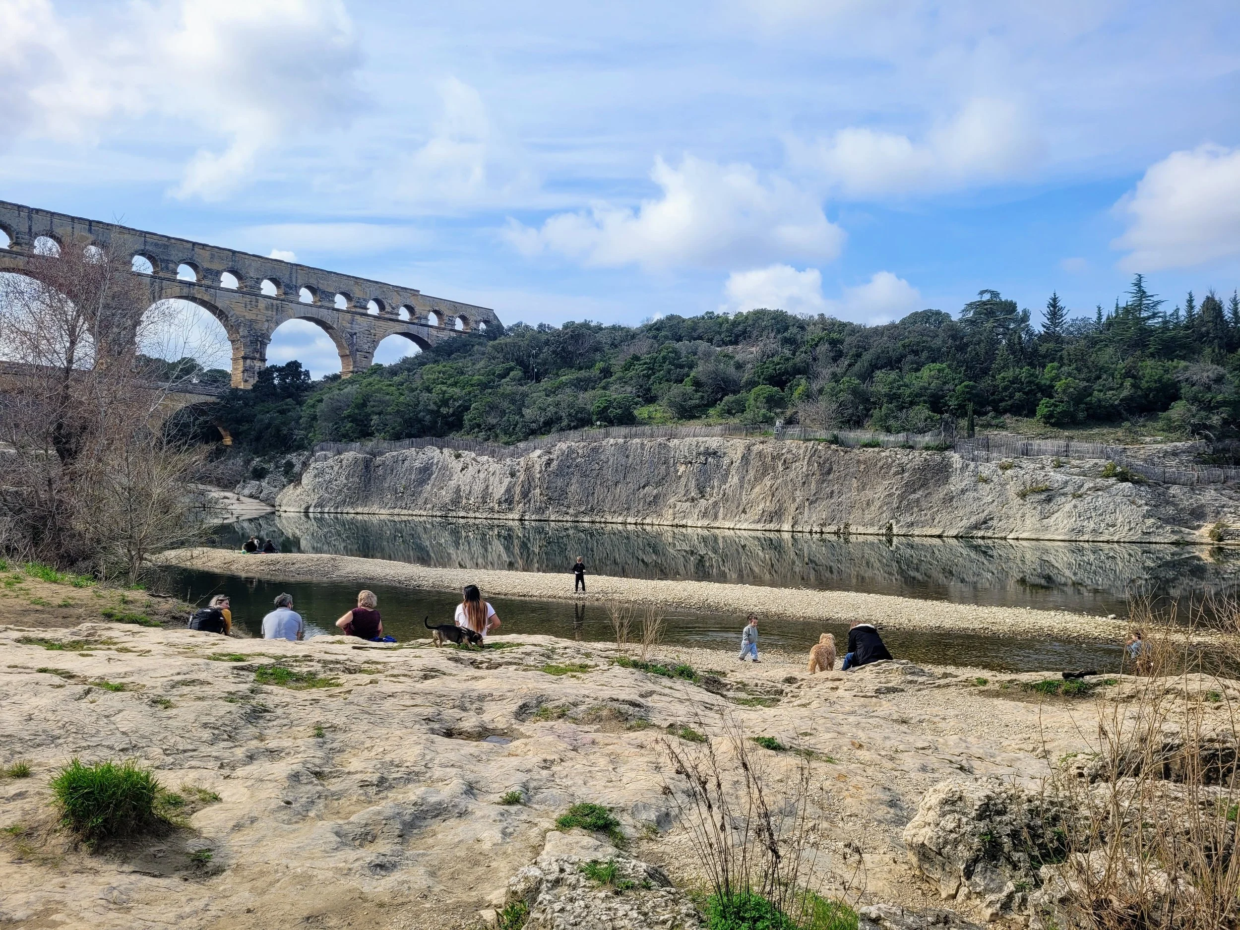 People sitting and walking along a rocky riverbank with a historic stone aqueduct and green hillside in the background under a partly cloudy sky.