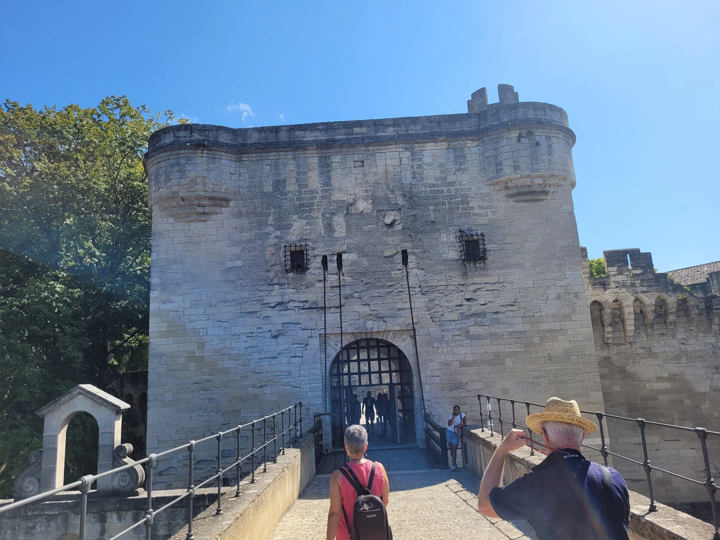 People entering a historic stone castle with a bridge and metal gates, surrounded by trees under a clear blue sky.