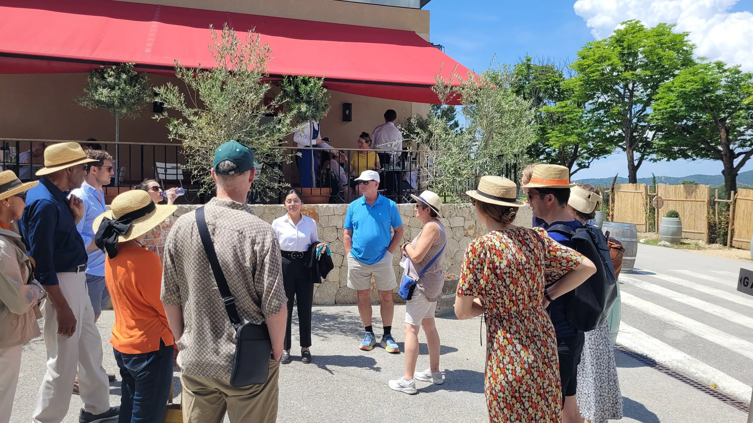 A group of people with summer hats gathered outside on a sunny day, facing a person who appears to be giving a tour or presentation. Some are standing, some are sitting, and others are on a raised outdoor patio with tables. There are trees, a red awning, and a partly cloudy sky in the background.