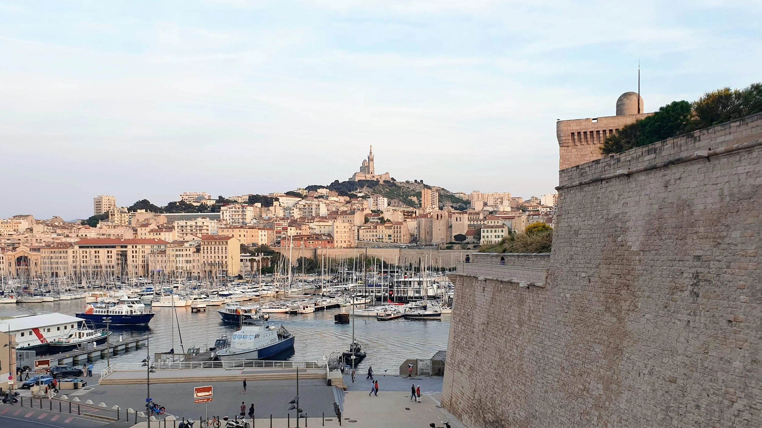 A cityscape featuring a marina with boats, a fortified wall, and a hill with numerous buildings and a prominent statue of the Virgin Mary at the top in the distance in Marseille