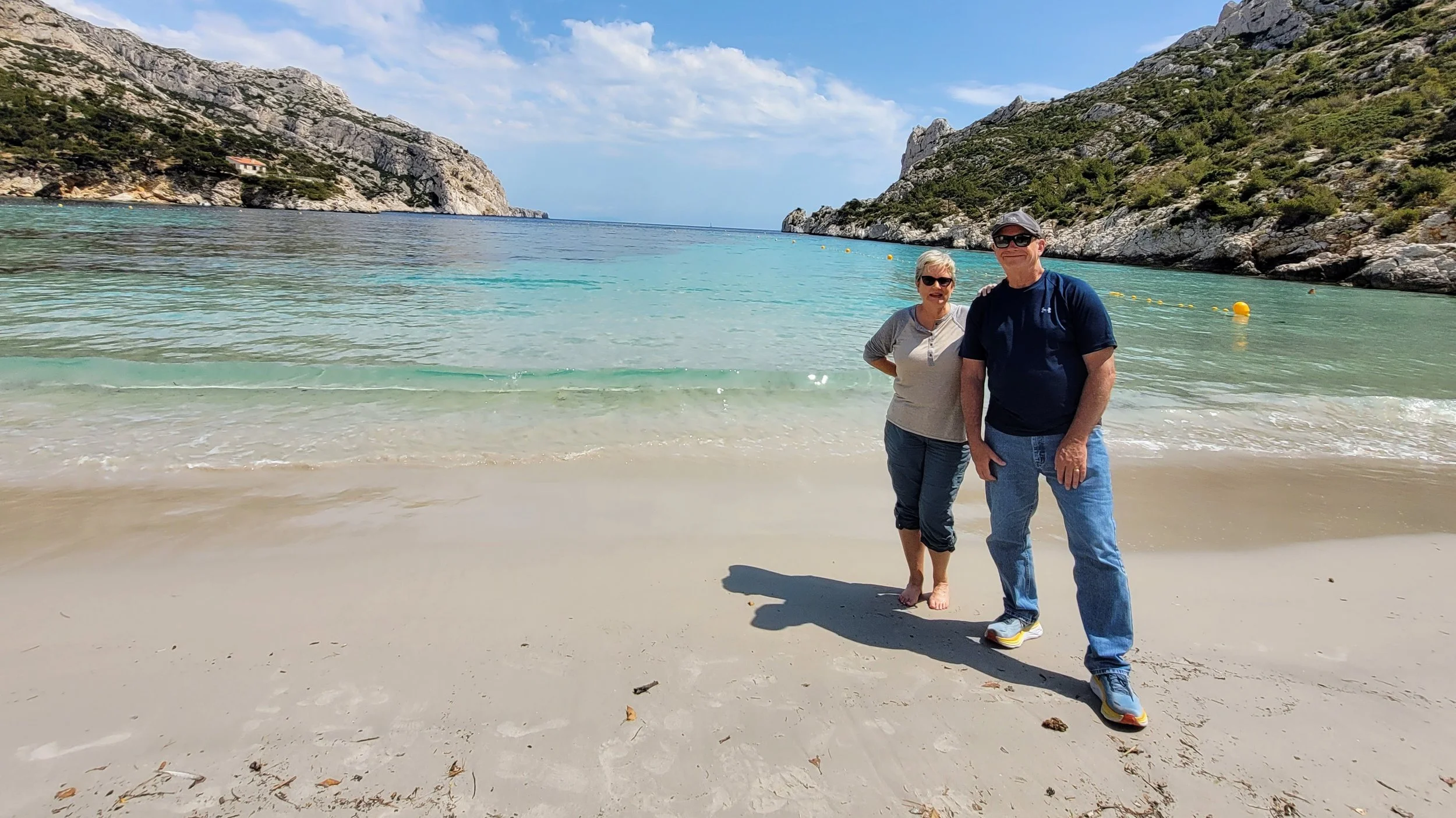 Two people walking on a sandy beach with clear blue water and rocky cliffs in the background during daytime, hidden gem near Marseille