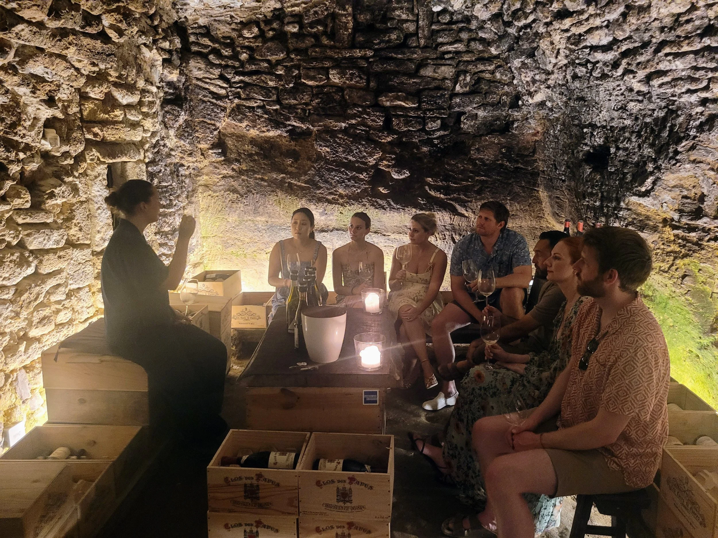 A group of people sitting around a woman who appears to be giving a wine tasting or conducting a presentation in a wine cellar with stone walls and warm lighting.