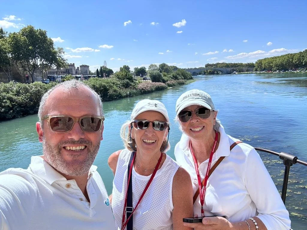 Three smiling people taking a selfie by a river on a sunny day.