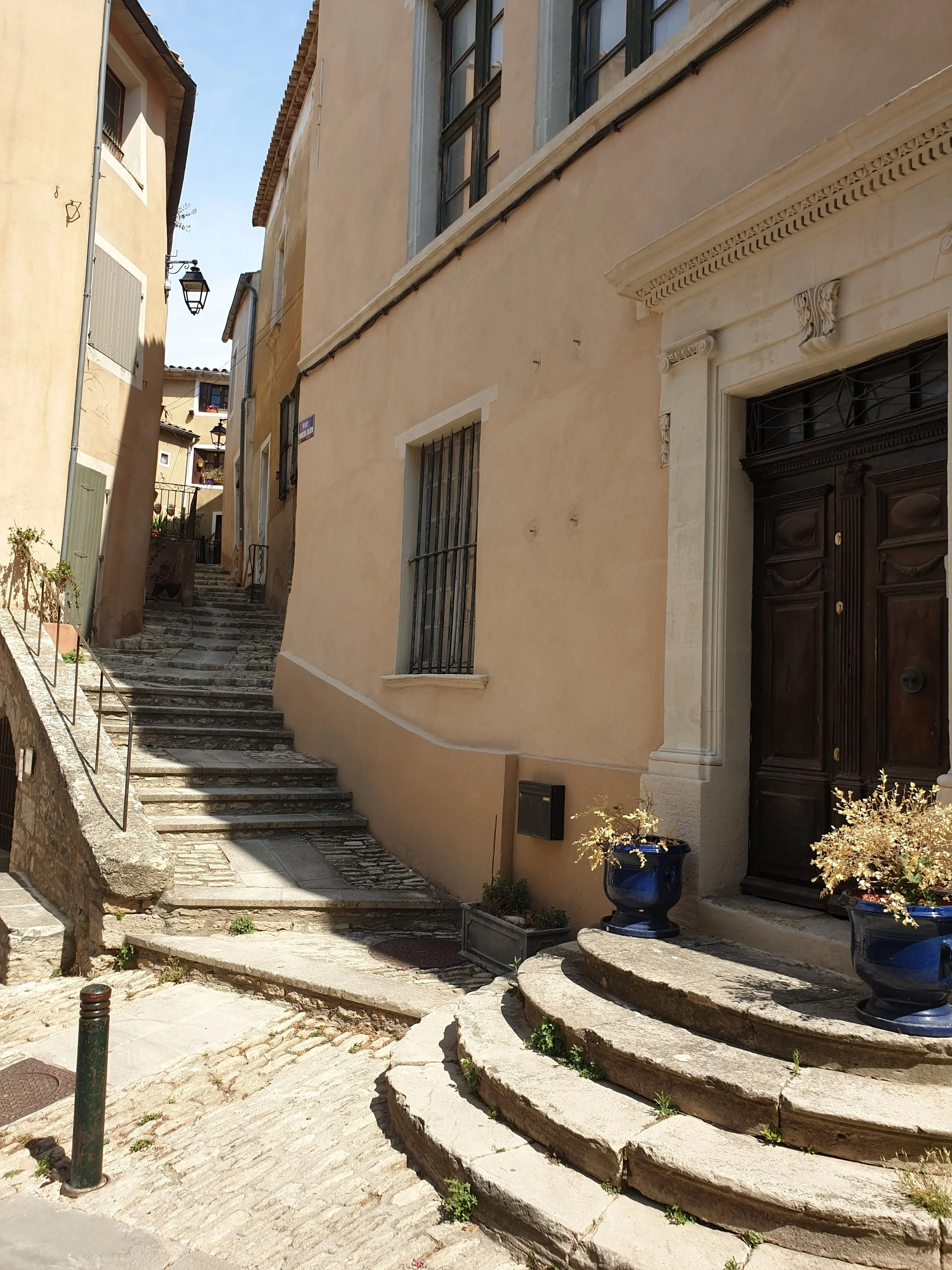 Narrow stone alleyway with stairs and buildings on both sides, potted plants near the entrance of a building, windows with iron bars, and a street lamp hanging from a building.