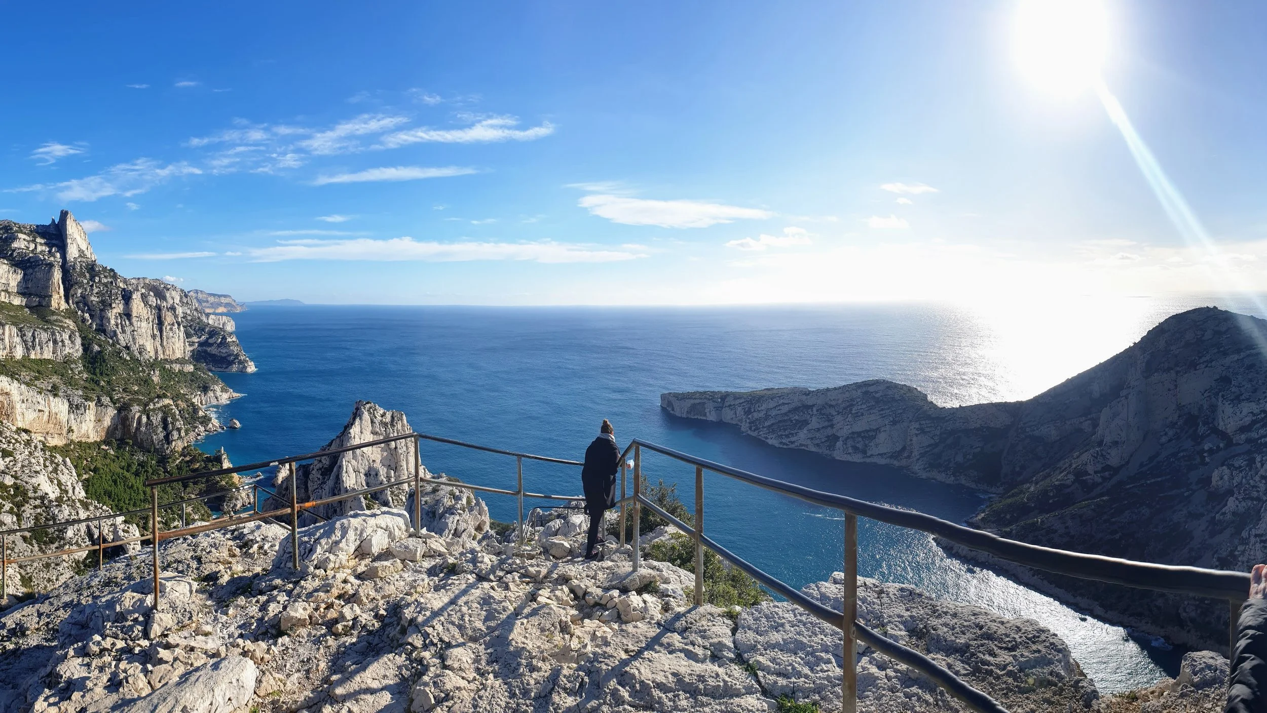 A person standing on a rocky cliff overlooking the ocean with mountains in the distance, a metal railing in the foreground, and a bright sun in the sky.