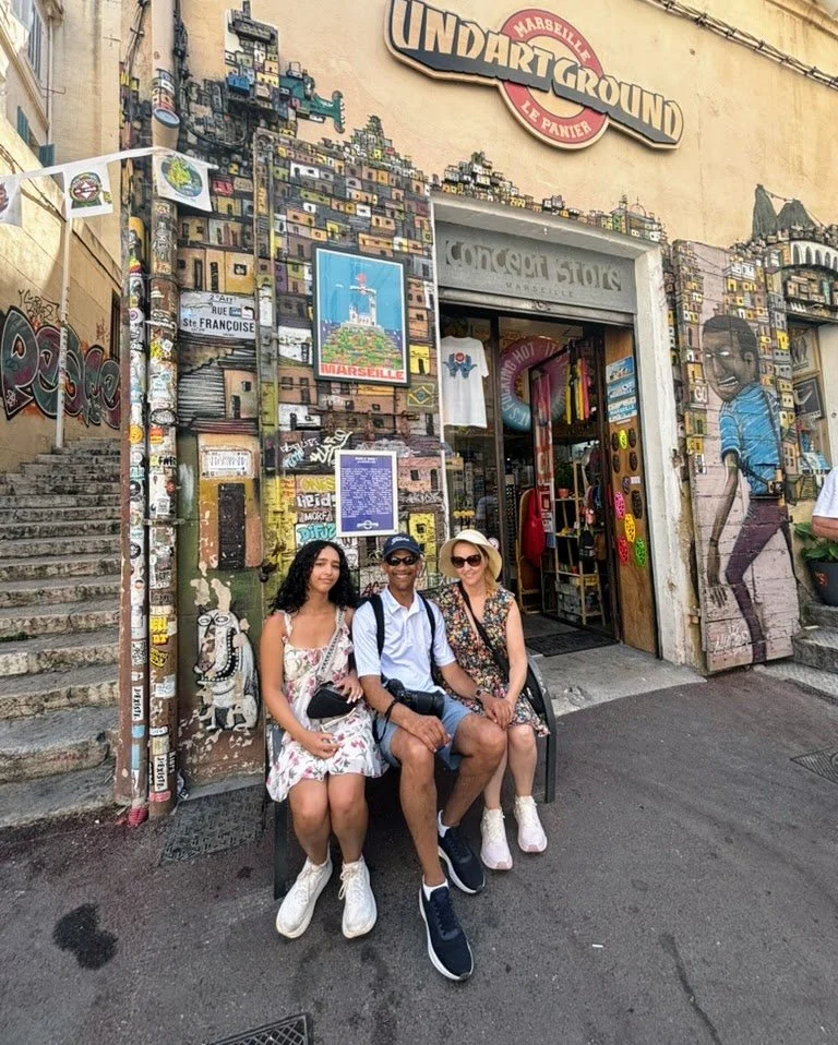 Three people sitting on a bench in front of a graffiti-covered building with a sign that reads 'Underground Marseilles.' The building entrance has colorful decorations and artwork, including a mural of a boy on the right side.