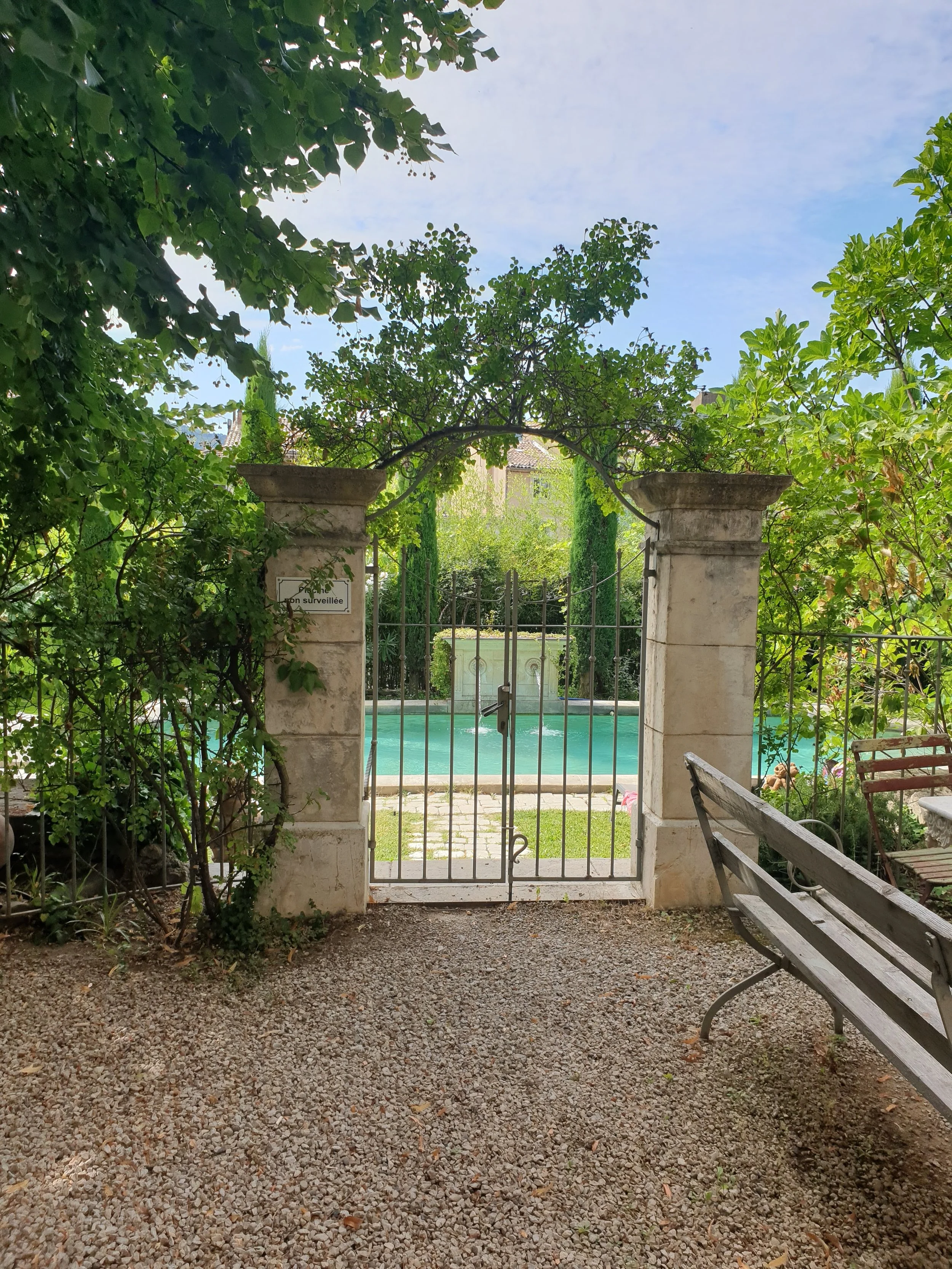 A view through a small wrought iron gate into a garden with a swimming pool, surrounded by plants and trees, with benches and a sign on the left pillar.