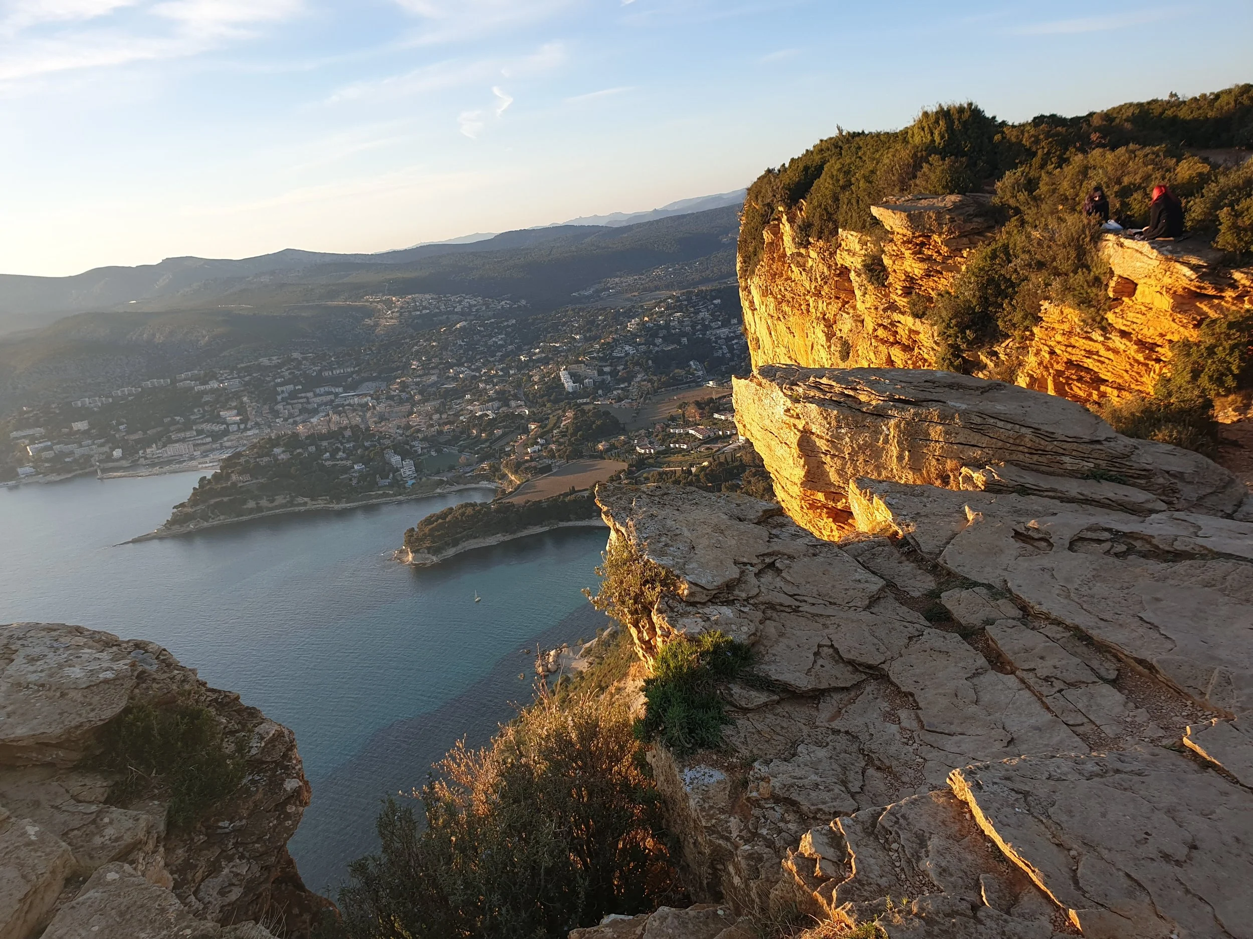 A scenic view from a rocky mountain ledge overlooking a bay in Cassis, with a city and hills in the background, during sunset.