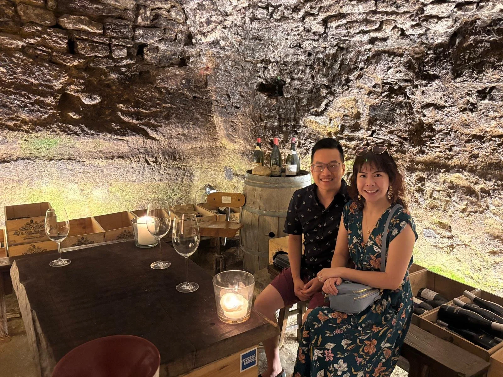 Two people, a man and a woman, smiling and sitting inside a wine cellar or tasting room with a stone ceiling. There are empty wine glasses on the table, a candle, and bottles of wine on a barrel behind them.
