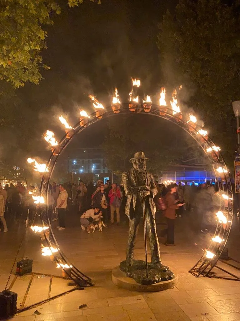 Statue of a man with a hat and cane under a circular metal arch with flames, in a crowded outdoor event at night.