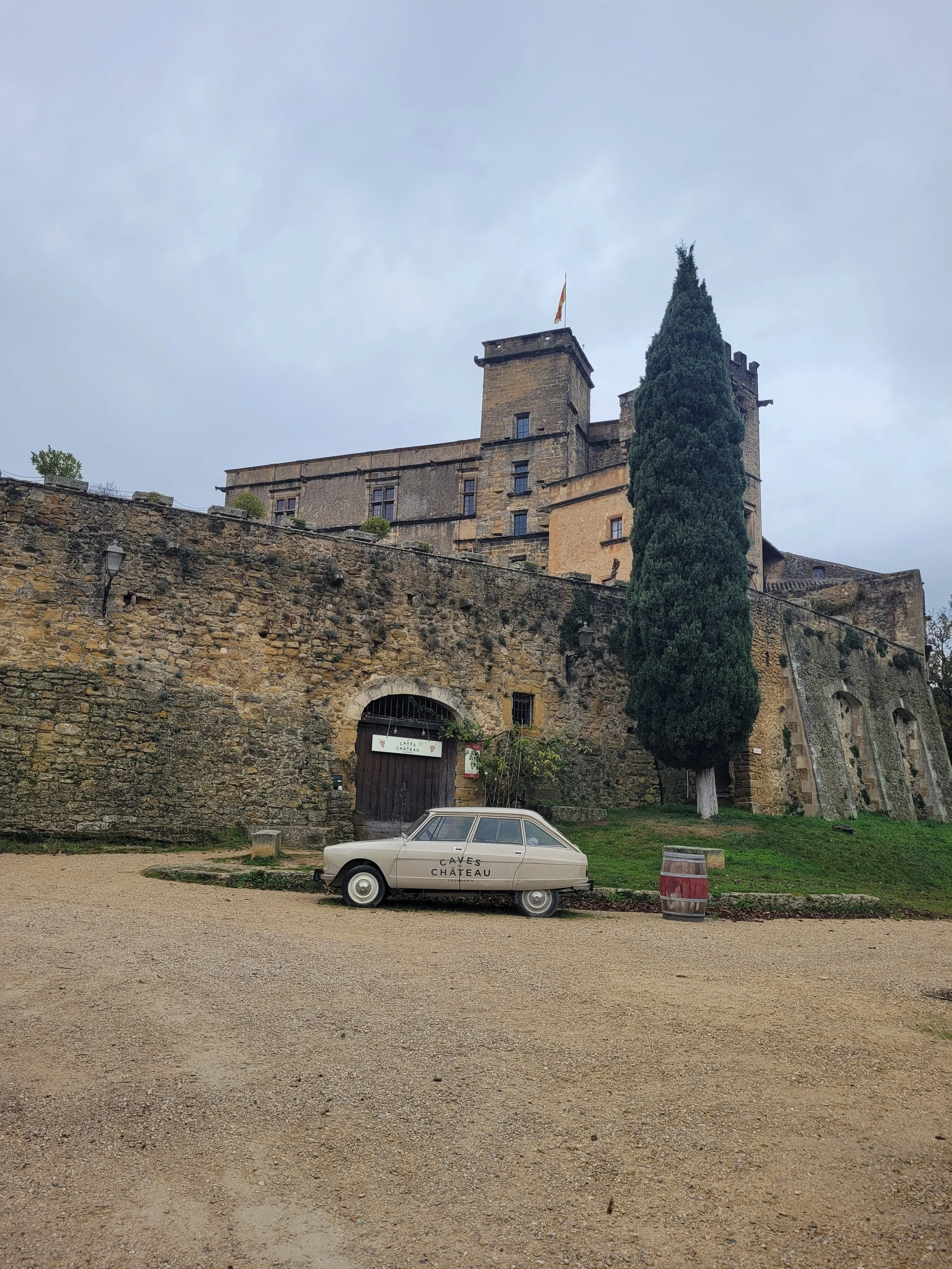 A castle on a hill with a large cypress tree in front, a vintage car with 'Caves Château' written on the side, and a wooden barrel to the right, under a cloudy sky.