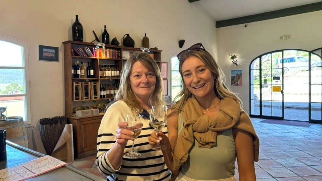 Two women smiling and holding wine glasses on a wine tasting day inside a tasting room with shelves of wine bottles behind them.